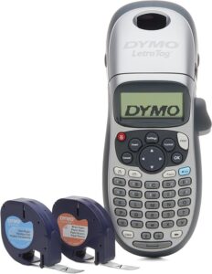 The image shows a DYMO LetraTag label maker. It has a compact, handheld design with a small screen and a full QWERTY keyboard. The label maker is silver and gray with various function buttons, including settings, insert, format, and print. The display shows the text 